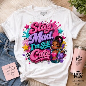 K&ouml;nnte beinhalten: Ein wei&szlig;es Kurzarm-T-Shirt mit einem Grafikdruck mit dem Text Stay Mad I'm Still Cute in Pink, Lila und T&uuml;rkis. Das Design zeigt eine Zeichentrickfigur mit dunkler Haut und lockigem Haar, umgeben von Sternen, Herzen und Farbspritzern.