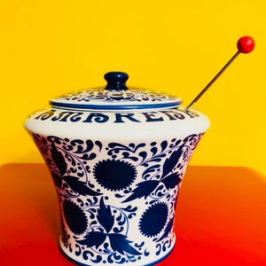May include: A white ceramic jar with a blue and white floral design. The jar has a lid and a red knob on the top. The jar is sitting on a red surface.