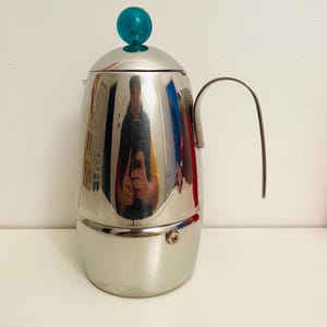 Pode incluir: Uma cafeteira italiana de aço inoxidável polido com um botão turquesa e uma alça curva. O metal reflete o ambiente. A cafeteira é projetada para preparar café expresso.
