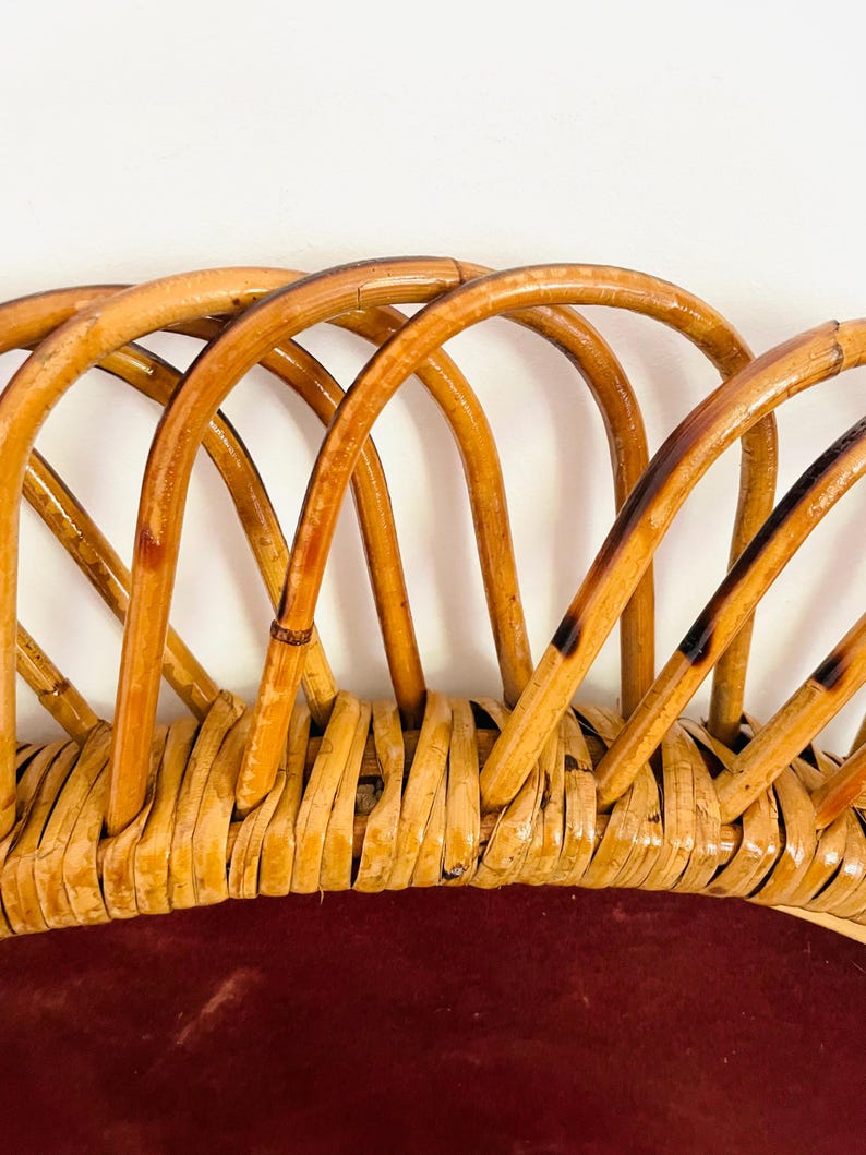 May include: Close-up of a vintage rattan chair back. The chair back features a woven base with a deep red cushion and arched rattan supports. The rattan is a light brown color with darker accents, creating a natural, textured look.