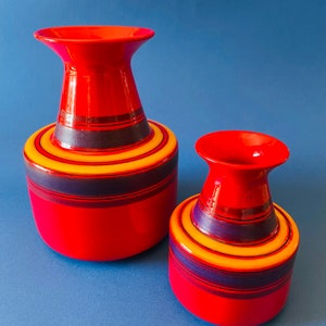 Op de afbeelding: Twee rode keramische vazen met gele, oranje en paarse strepen. De grotere vaas is hoger en breder dan de kleinere vaas.