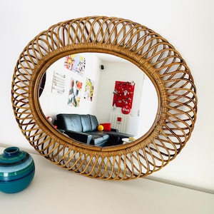 May include: Oval rattan mirror with a woven design. The mirror has a natural brown color and reflects a room with a black sofa and colorful artwork. A teal and blue vase sits on the white surface below the mirror.
