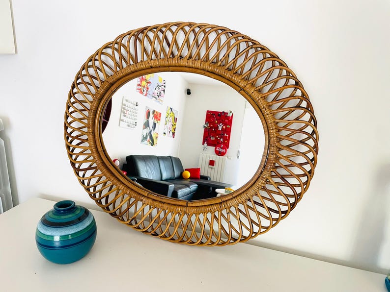 May include: Oval rattan mirror with a woven design. The mirror has a natural brown color and reflects a room with a black sofa and colorful artwork. A teal and blue vase sits on the white surface below the mirror.