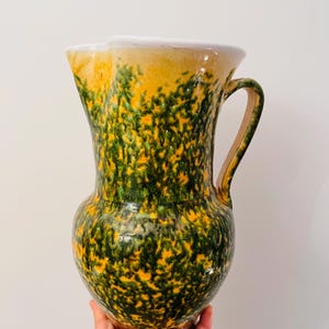 Peut inclure: Une grande cruche en céramique avec une large ouverture et une anse. La cruche est principalement jaune avec un motif vert tacheté et une base marron. Le bord est blanc.