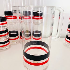 May include: A set of clear acrylic glasses and a pitcher with black and red stripes. The glasses are stacked and the pitcher has a handle.