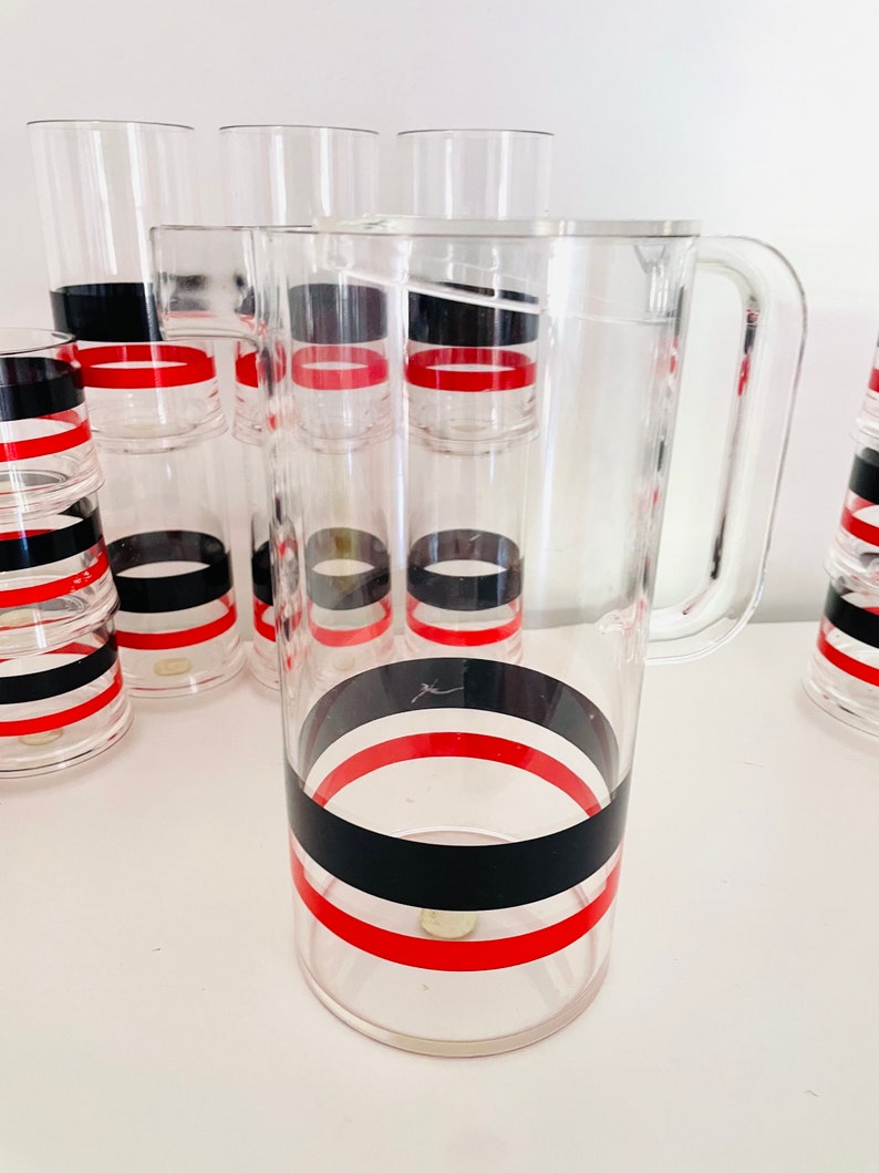 May include: A set of clear acrylic glasses and a pitcher with black and red stripes. The glasses are stacked and the pitcher has a handle.
