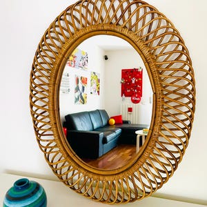 May include: Oval rattan mirror with a woven frame. The mirror reflects a room with a black sofa, artwork, and a blue and green vase. The frame is a warm brown color.