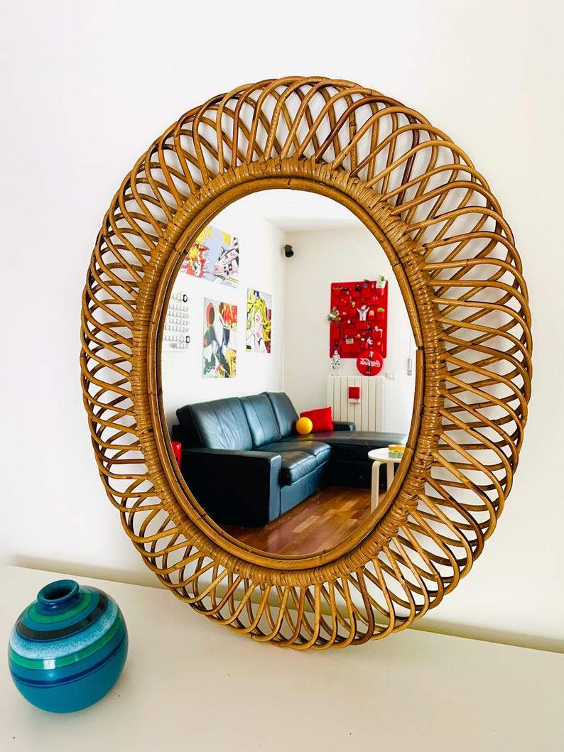 May include: Oval rattan mirror with a woven frame. The mirror reflects a room with a black sofa, artwork, and a blue and green vase. The frame is a warm brown color.