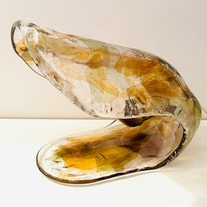 Może przedstawiać: Unikalna, abstrakcyjna lampa szklana o falistym, organicznym kształcie. Lampa wykonana jest z przezroczystego szkła z wtopionymi kawałkami szkła w kolorze złotym, brązowym i zielonym. Lampa jest oświetlona żar&oacute;wką umieszczoną w podstawie.