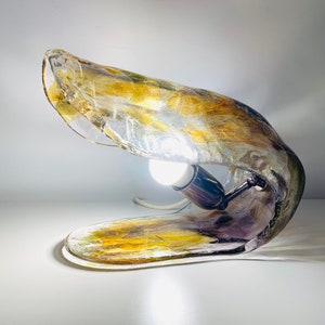 Może przedstawiać: Unikalna lampa stołowa wykonana z przezroczystego szkła z złotymi i fioletowymi akcentami. Lampa ma zakrzywiony, organiczny kształt, a wewnątrz widoczna jest żar&oacute;wka.