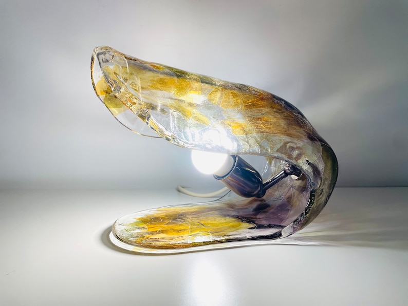 Może przedstawiać: Unikalna lampa stołowa wykonana z przezroczystego szkła z złotymi i fioletowymi akcentami. Lampa ma zakrzywiony, organiczny kształt, a wewnątrz widoczna jest żar&oacute;wka.