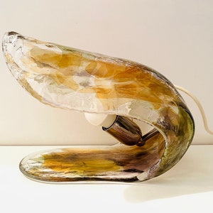 Może przedstawiać: Unikalna lampa stołowa z zakrzywioną, bursztynową podstawą ze szkła. Podstawa ma wirujący, marmurowy wz&oacute;r i przezroczysty element ze szkła. Lampa ma biały przew&oacute;d i białą wtyczkę.