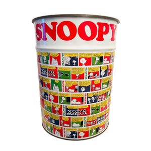 May include: A cylindrical metal container with a white background and a red "SNOOPY" logo. The container is decorated with colourful comic strips featuring Peanuts characters. The rim is a dark brown colour.