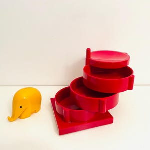 May include: A red plastic tiered jewelry organizer with four levels. The organizer is shaped like a spiral staircase. A small yellow plastic elephant figurine is in the foreground.