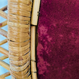 May include: Close-up of a section of a rattan chair. The frame is made of woven, light brown rattan, with a deep burgundy fabric visible in the background. The chair's design features a combination of woven and solid rattan elements.