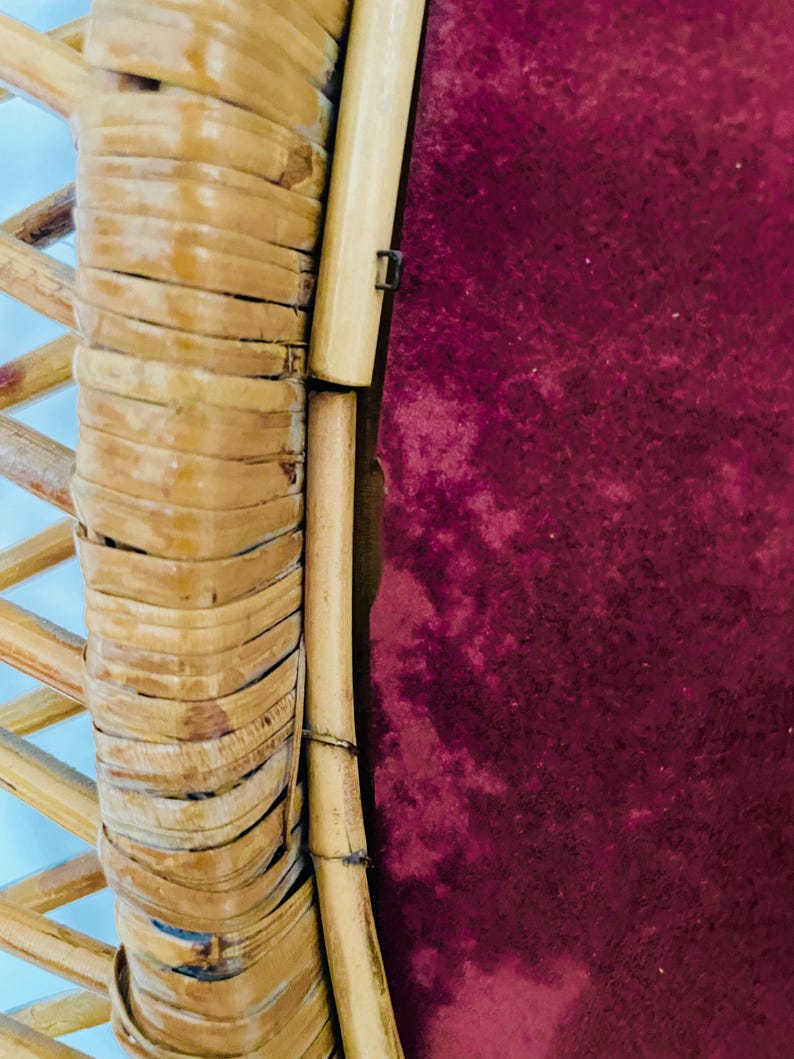 May include: Close-up of a section of a rattan chair. The frame is made of woven, light brown rattan, with a deep burgundy fabric visible in the background. The chair's design features a combination of woven and solid rattan elements.