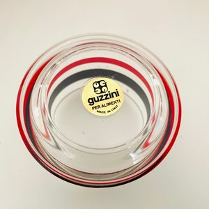 May include: A clear glass bowl with red and black stripes around the rim. The bowl has a gold sticker on the bottom that says "Guzzini Per Alimenti Made in Italy".