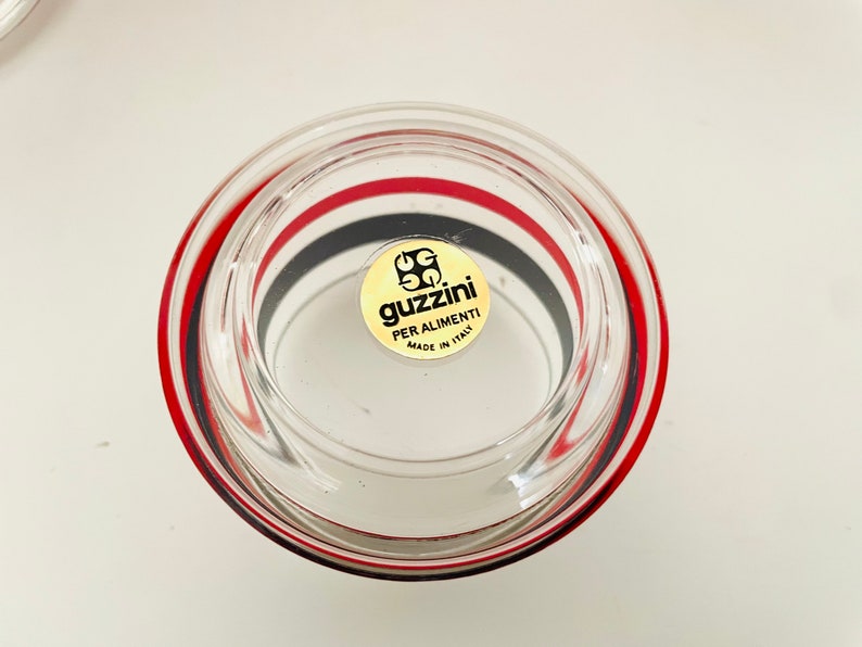 May include: A clear glass bowl with red and black stripes around the rim. The bowl has a gold sticker on the bottom that says "Guzzini Per Alimenti Made in Italy".