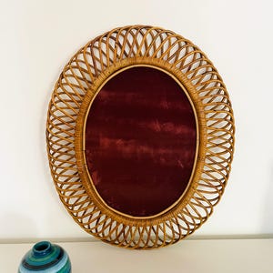 May include: Oval rattan mirror with a deep red velvet center. The mirror is surrounded by a woven rattan frame. A small, blue and green striped ceramic vase sits below the mirror.