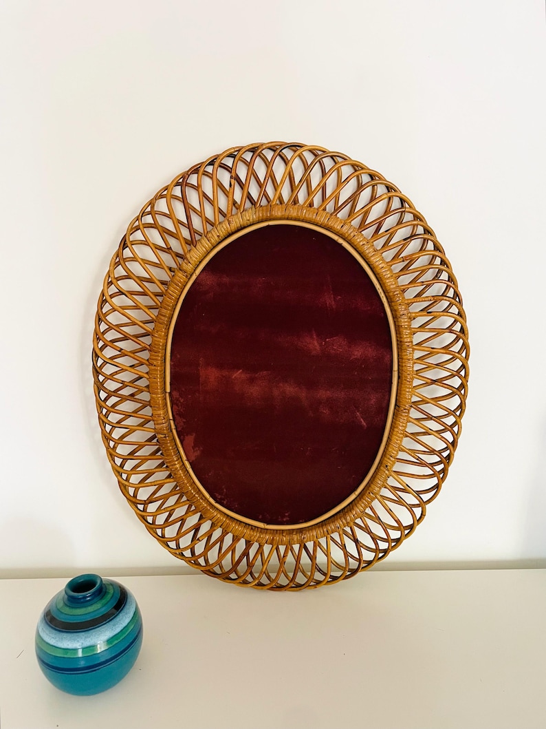 May include: Oval rattan mirror with a deep red velvet center. The mirror is surrounded by a woven rattan frame. A small, blue and green striped ceramic vase sits below the mirror.