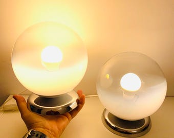 May include: Two white glass globe table lamps with chrome bases. The lamps are turned on and the light bulbs are visible.