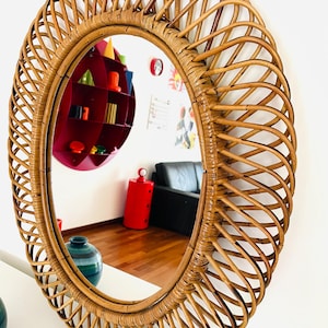 May include: Oval-shaped rattan mirror with a woven design. The mirror reflects a room with a red shelf, a black couch, and a wooden floor. The frame is a warm brown color.