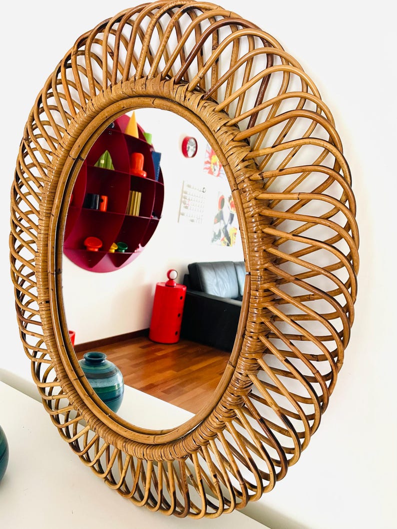 May include: Oval-shaped rattan mirror with a woven design. The mirror reflects a room with a red shelf, a black couch, and a wooden floor. The frame is a warm brown color.