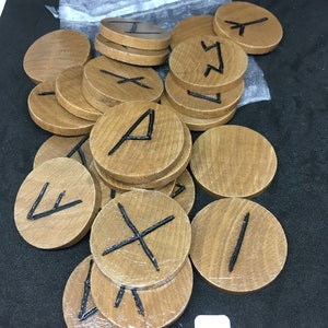 May include: A set of wooden runes with black painted symbols. The runes are circular and have a natural wood grain finish. The runes are scattered on a black surface.