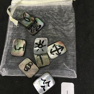 Puede incluir: Un conjunto de ocho fichas de concha negra y blanca con diferentes símbolos y diseños. Las fichas están en una bolsa de malla blanca. Las fichas están numeradas del 1 al 8.