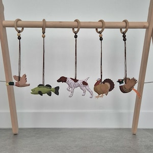 Può includere: Una palestrina in legno per bambini con giocattoli di animali in feltro appesi. I giocattoli includono un'anatra, un pesce, un cane, un tacchino e un fagiano. I giocattoli sono sospesi ad anelli di legno con cordoncini marroni.