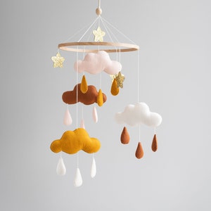 May include: A wooden baby mobile with felt clouds, stars, and raindrops in shades of pink, brown, white, and gold. The mobile is hanging from a white ceiling.