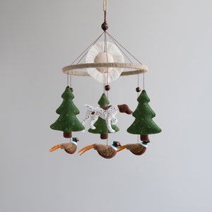 May include: A woodland-themed baby mobile with a white and brown spotted dog, three green trees, and three brown and orange pheasants. The mobile is suspended from a brown wooden ring with a white and brown sun-shaped decoration.