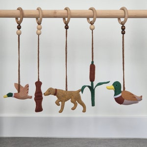 May include: A wooden mobile with five felt ornaments hanging from it. The ornaments include a brown and white duck, a brown dog, a brown and white bird, a brown tube, and a green reed.
