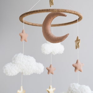 Op de afbeelding: Een babymobiel met een bruine vilt halve maan, witte pluche wolken en gouden en bruine vilt sterren. De mobiel hangt aan een houten ring.