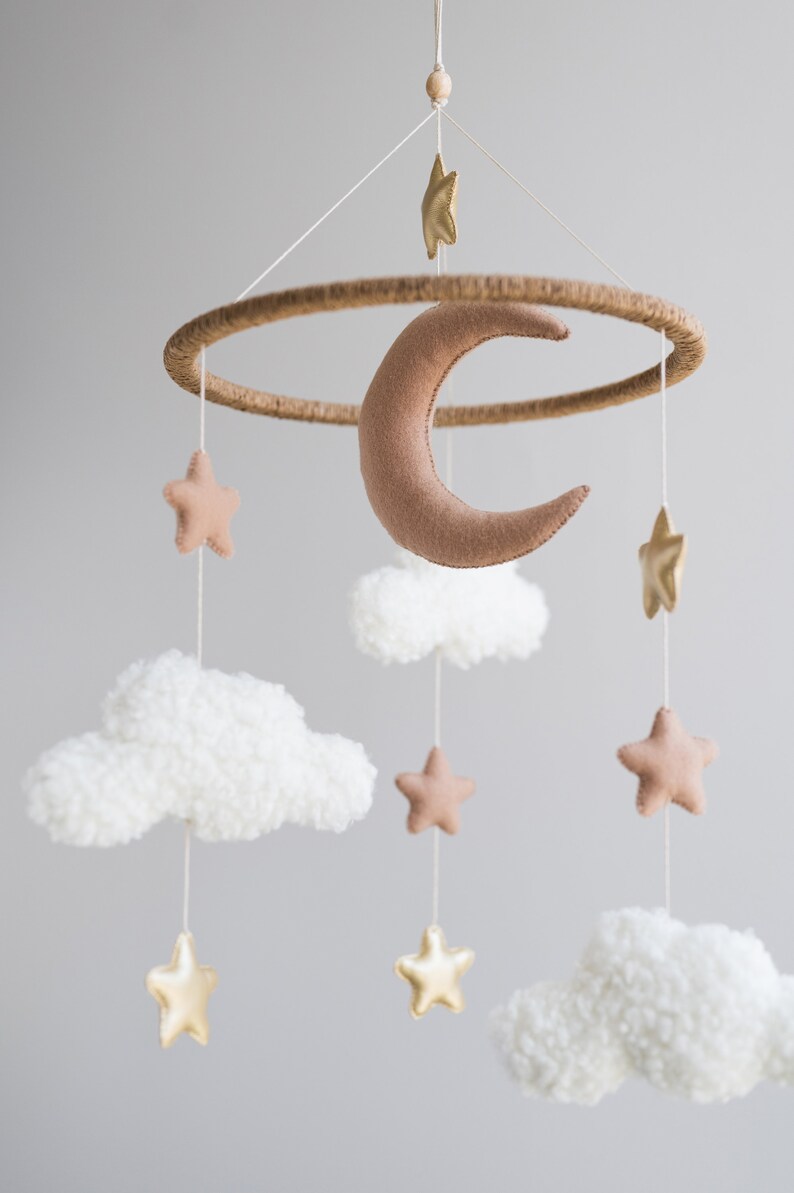 Op de afbeelding: Een babymobiel met een bruine vilt halve maan, witte pluche wolken en gouden en bruine vilt sterren. De mobiel hangt aan een houten ring.