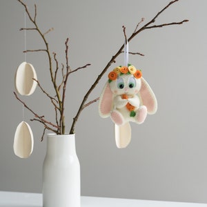 Op de afbeelding: Een witte keramische vaas bevat kale takken met hangende houten eierornamenten. Een vilten konijnenornament met een bloemenkrans en wortel zit op een tak, wat een lente- of Paasthema creëert.