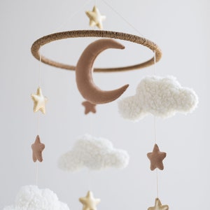 Op de afbeelding: Een witte, bruine en gouden babymobiel met een halve maan, wolken en sterren. De mobiel is gemaakt van vilt en hangt aan een houten ring.