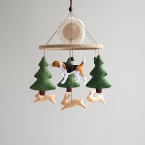 May include: A felt baby mobile with a brown and white dog, three brown bunnies, and two green trees hanging from a wooden ring. The mobile is suspended from a white ceiling.