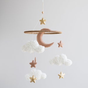 Op de afbeelding: Een witte en bruine babymobiel met een halve maan, sterren en pluche wolken. De mobiel is gemaakt van vilt en hangt aan een houten ring.