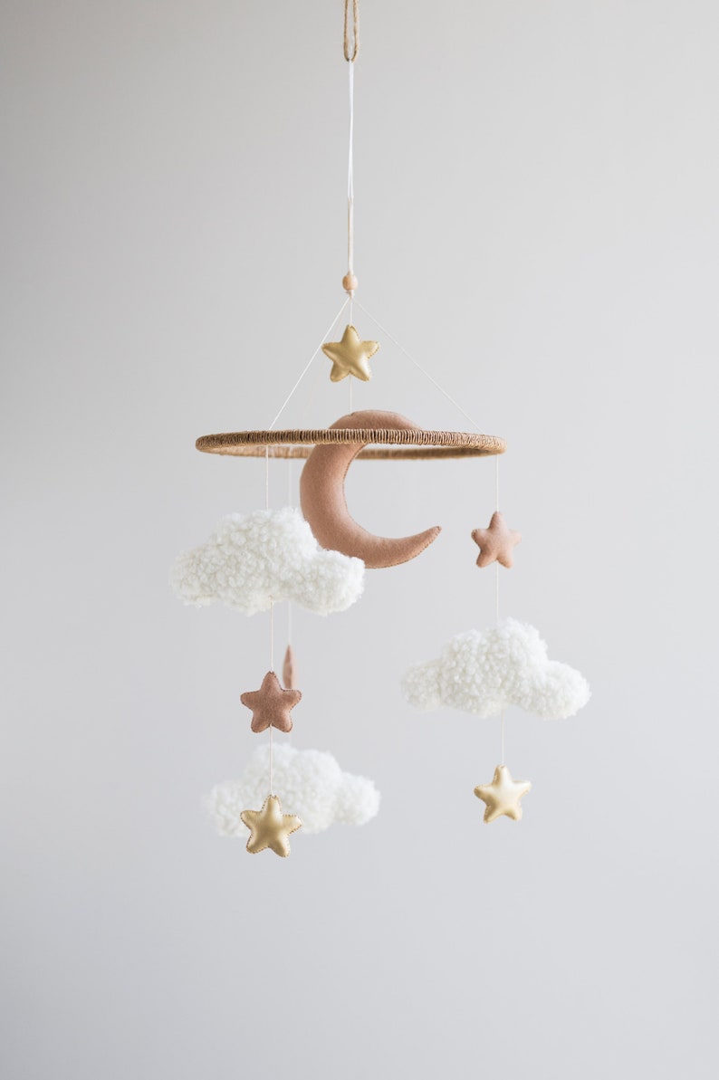 Op de afbeelding: Een witte en bruine babymobiel met een halve maan, sterren en pluche wolken. De mobiel is gemaakt van vilt en hangt aan een houten ring.