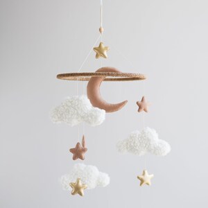 Op de afbeelding: Een witte en bruine babymobiel met een halve maan, sterren en pluche wolken. De mobiel is gemaakt van vilt en hangt aan een houten ring.
