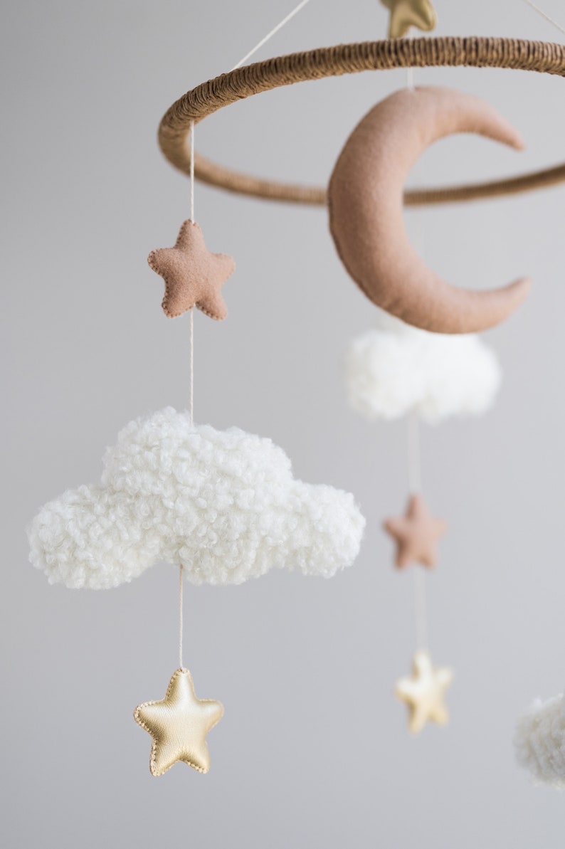 Op de afbeelding: Een mobiel met een witte, pluche wolk, een bruine vilt halve maan en gouden en bruine vilt sterren die aan een houten ring hangen. Deze mobiel is perfect voor een babykamer.
