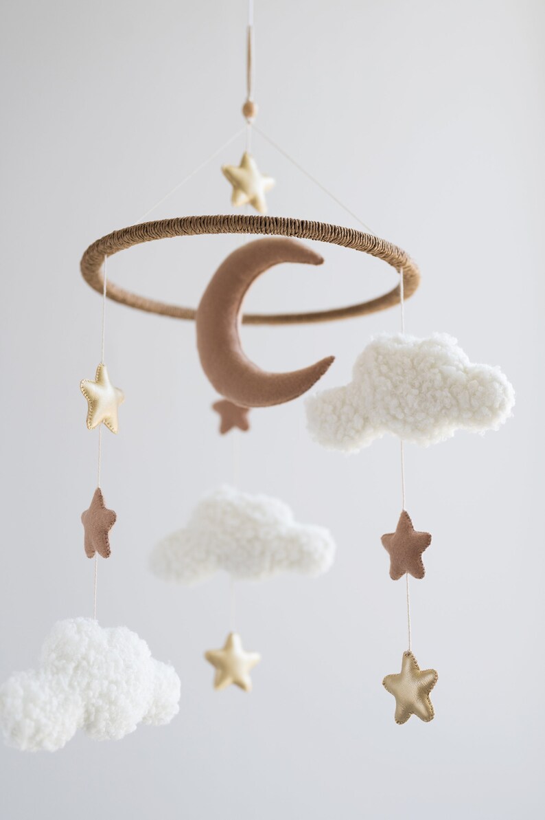 Op de afbeelding: Een witte, bruine en gouden babymobiel met een halve maan, wolken en sterren. De mobiel is gemaakt van vilt en hangt aan een houten ring.