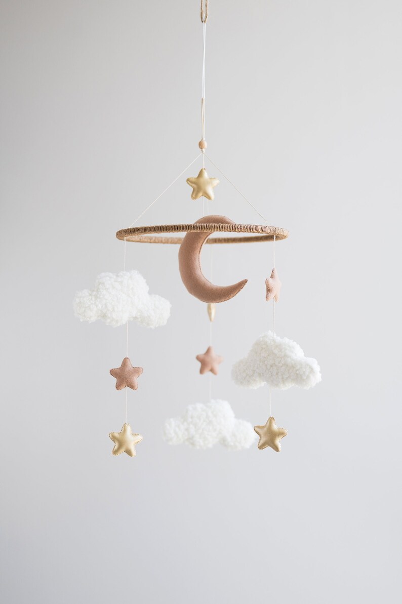 Op de afbeelding: Een witte en gouden babymobiel met een halve maan, sterren en pluche wolken. De mobiel hangt aan een wit touw.