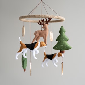 Op de afbeelding: Een houten mobiel met vilten bosdieren, waaronder een hert, twee honden en een groene boom. De mobiel hangt aan een touw en heeft houten kralen aan de bovenkant.