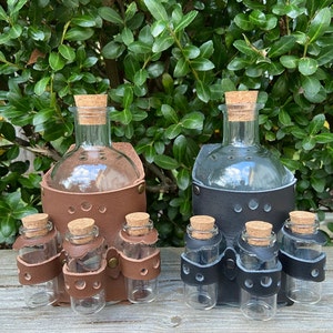 May include: Two leather-wrapped glass bottles with cork stoppers. Each bottle has three smaller glass vials attached to the side. One bottle is brown leather and the other is black leather.