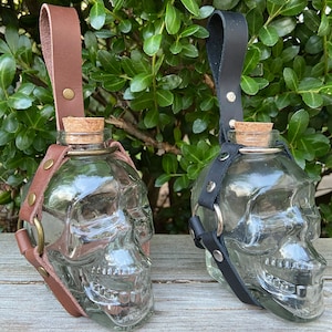 May include: Two skull-shaped glass bottles with cork stoppers. One has a brown leather strap and the other has a black leather strap. Both bottles have a clear, transparent appearance and are displayed against a backdrop of green foliage.
