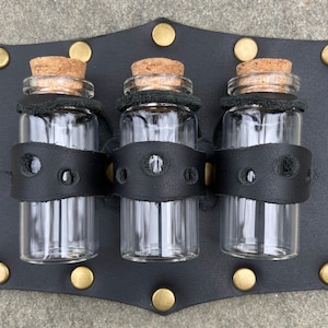 May include: Three glass bottles with cork tops are held in a black leather holder with metal rivets. The bottles are clear and have a black leather band around the middle.
