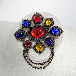 Könnte beinhalten: Eine Vintage-Brosche mit einem Sternburst-Design mit roten, blauen und gelben Glassteinen. Die Brosche hat eine baumelnde Kette.