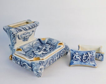 Op de afbeelding: Een blauwe en witte asbak van keramiek met een gouden rand en een bijpassende kleine doos. De asbak heeft een ontwerp van cherubijnen in een tuinsetting.
